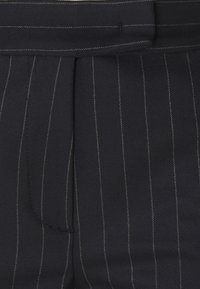 Tissu à fines rayures foncées avec de fines rayures blanches verticales et une taille repliée, montrant un gros plan de pantalons sur mesure.