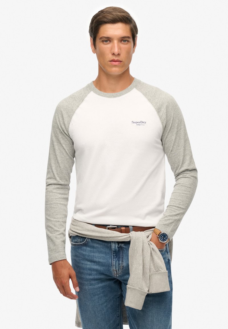 Biała koszulka z długim rękawem z szarymi rękawami raglanowymi, z materiału bawełnianego, z małym logo na piersi. Noszona z dżinsami i swetrem związanym w pasie.