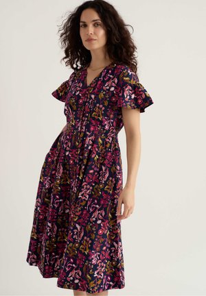 Kobieta o kręconych włosach, ubrana w granatową sukienkę z różowymi, czerwonymi i żółtymi kwiatowymi wzorami oraz rozkloszowanymi rękawami, stojąca na jednolitym tle.