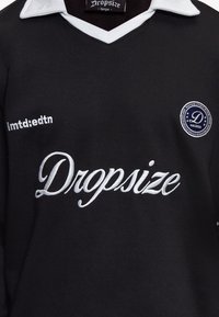 Chemise noire avec col blanc ornée d'un texte brodé blanc "Dropsize" sur la poitrine, petit logo circulaire, et texte "lmtd:edtn" sur le côté gauche.