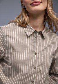 Chemise beige à boutons avec des rayures blanches verticales. Elle comporte un col, une poche avant et une texture lisse. Détails de design subtils et discrets.