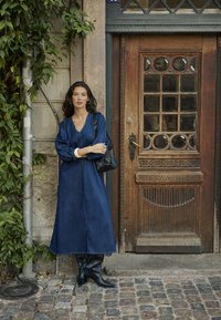 Denim maksinska obleka z V-izrezom, napihnjenimi rokavi in stranskim razporkom, v kombinaciji z črnými škornji do kolen in črno torbico. Ozadje prikazuje lesenih vrata.