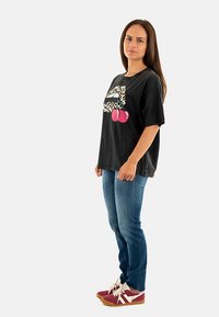Zwart grafisch T-shirt met luipaardprintlippen en roze kersen, gecombineerd met blauwe spijkerbroek en bordeauxrode sneakers met witte accenten.