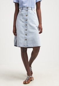 Hellblauer Jeansrock mit tailliertem Bund, A-Linien-Form, vorderer Knopfverschluss und seitlichen Taschen. Begleitet von verzierten Sandalen.