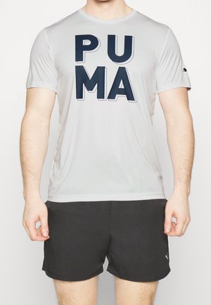 Mies, joka pukeutunut vaaleanharmaaseen Puma T-paitaan, jossa isot tummansiniset kirjaimet, ja mustiin Puma-shortseihin, seisoo yksivärisen taustan edessä.