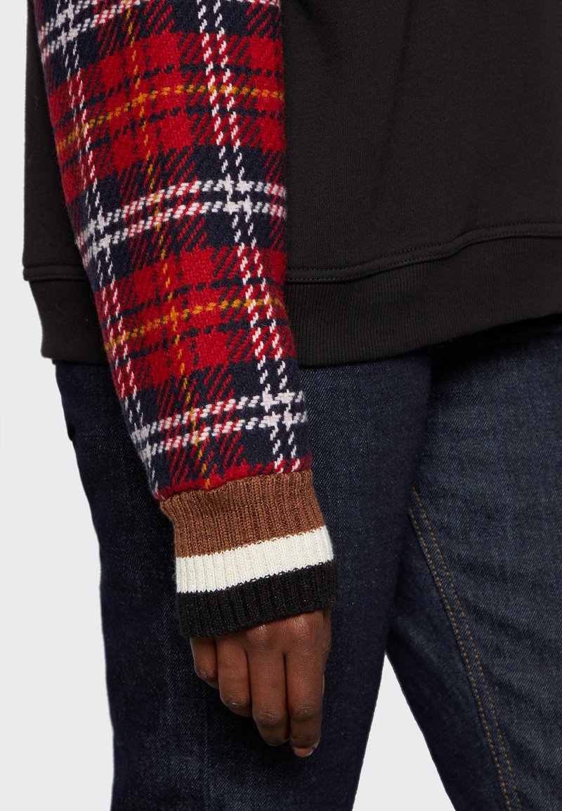 Czarna bluza z rękawem w kratę w kolorze czerwonym, granatowym i białym; prążkowany brązowy i biały mankiet; widoczne ciemne dżinsy.