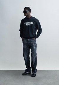 Black sweatshirt with "DSQUARED2 MILANO" in white, paired with faded blue jeans and black footwear. Model wears dark sunglasses.