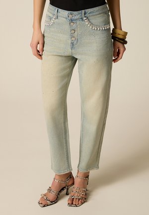 Jeans a lavaggio chiaro con dettagli di perle e perline sulle tasche e sulla parte superiore delle gambe, chiusura con bottoni, indossati con top nero e sandali con cinturini in pelle di serpente.