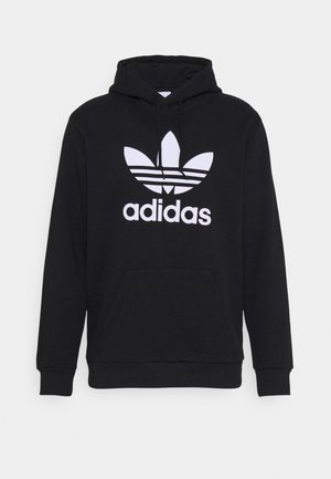 Personne portant une veste polaire color block, un sweat à capuche noir Adidas, un pantalon tie-dye blanc et noir et des baskets New Balance grises, debout les mains dans les poches.