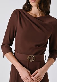 Donna con un abito lungo a maniche di colore marrone, con una cintura larga che presenta una fibbia rotonda dorata, le mani incrociate in vita.