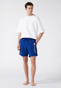 T-shirt blanche surdimensionnée, à manches courtes, associée à un short de bain bleu avec une taille élastique et un logo sur le côté.