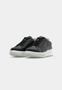 Baskets noires montantes avec lacets noirs, côtés à motifs, semelles blanches rembourrées, et nom de la marque embossé sur la semelle et la languette.