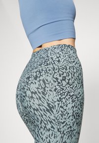 Gros plan d'une personne portant un legging bleu à motifs taille haute et un crop top bleu clair, montrant le détail de la poche latérale.
