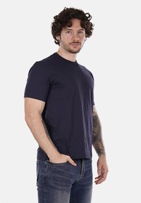 T-shirt in cotone blu navy con maniche corte, colletto rotondo, tessuto leggero e un sottile dettaglio del logo sul petto. Indossato con jeans.