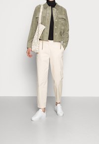 Olive green suede jacket with front pockets, black turtleneck, cream trousers, and white sneakers; carrying a patterned beige shoulder bag.
