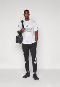 Άνδρας με λευκό μπλουζάκι Adidas, μαύρο παντελόνι φόρμας Adidas, μαύρα αθλητικά παπούτσια, κρατώντας μια μαύρη τσάντα ώμου, στέκεται σε άσπρο φόντο.