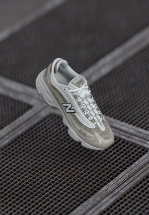Hvid og lysegrå New Balance 1000 sneaker, der svæver over en gulv med metalnet med diagonalt krydsende linjer.