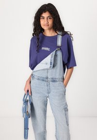 Salopette en denim avec jambes fuselées et bretelles réglables, associée à un t-shirt à manches courtes violet avec un texte blanc. Petit sac à main bleu.