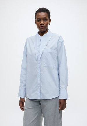 Gaiši zils krekls ar pogām, augstu apkakli, garām piedurknēm un krūšu kabatu. Gluda, viegla auduma ar modernu griezumu.