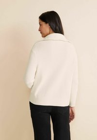 Pull en tricot crème avec un col châle, texture côtelée, épaules tombantes et coupe décontractée, associé à un pantalon noir.