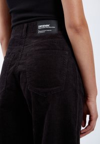Pantalones de terciopelo negro con gruesas estrías verticales. Presentan bolsillos traseros, una etiqueta negra en la cinturilla y un corte relajado.