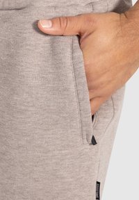 Pantalones de chándal grises claros con una tela suave y texturizada. Bolsillo lateral visible, mano descansando dentro. Etiqueta negra en la costura lateral.