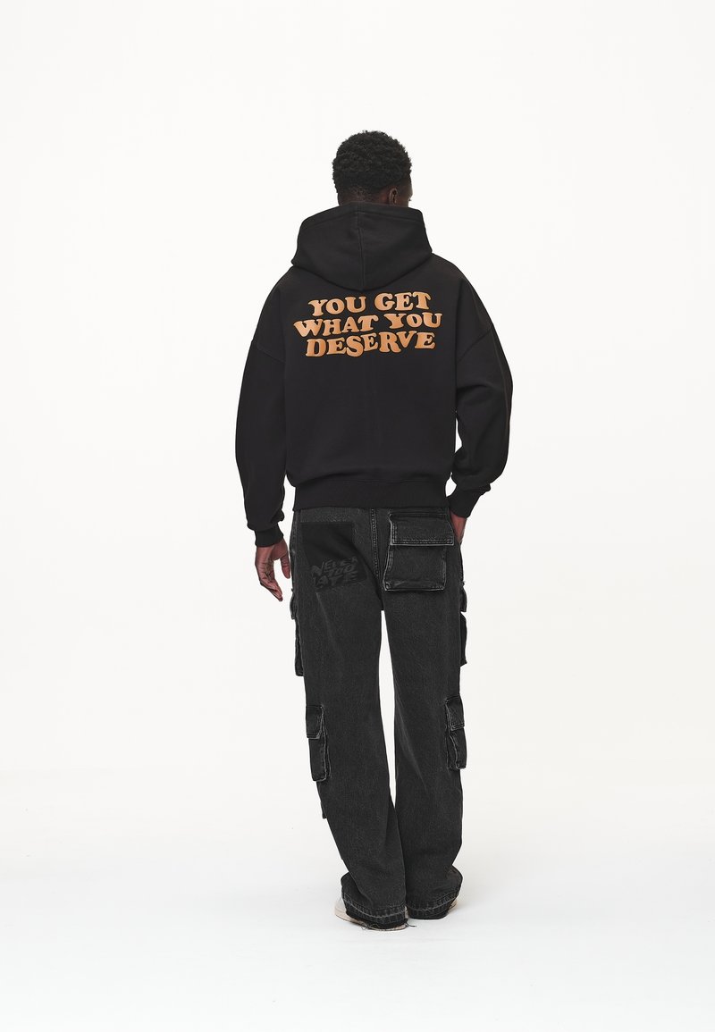 Personne portant un sweat à capuche noir avec le texte orange « YOU GET WHAT YOU DESERVE » dans le dos, associé à un pantalon cargo noir et des baskets blanches.
