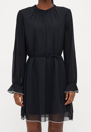 Femme portant une robe noire mi-longue avec des manches longues transparentes, des poignets plissés, une ceinture nouée à la taille et un col rond avec un bouton.