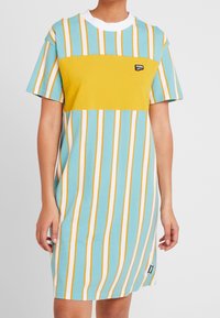 T-shirtklänning i ljusblått med gula och vita vertikala ränder. Har en gul blockpanel och logotyp på bröstet. Kort ärm.