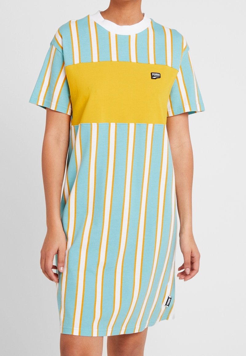 T-shirtklänning i ljusblått med gula och vita vertikala ränder. Har en gul blockpanel och logotyp på bröstet. Kort ärm.