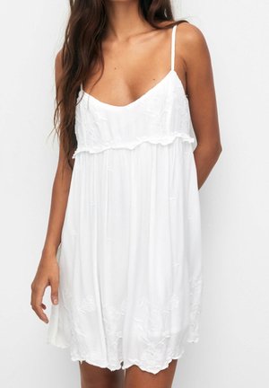 Woman wearing a sleeveless white dress with thin straps and delicate floral embroidery, standing against a plain white background.