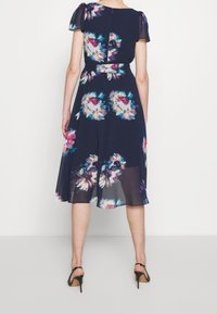 Kukallinen mekko tummansinisestä kankaasta, jossa on puoliksi läpinäkyvät lyhyet hihat. Mekon vyötärö on istuva ja helma virtaa, koristeltuna värikkäillä kukkakuoseilla.