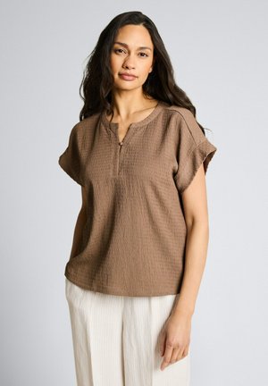 Vrouw met lang donker haar, die een kort mouw bruin textuur top en lichte gestreepte broek draagt, staand tegen een effen achtergrond.