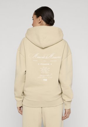 Femme portant un sweat à capuche beige avec le texte blanc « Moments to Memories » et les détails de la marque au dos, debout dos à la caméra.
