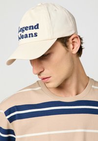 Gorra de béisbol beige con el texto bordado en azul "Legend Jeans". Llevada con una camiseta de rayas beige que presenta rayas azules y blancas.