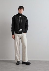 Chaqueta bomber negra sobre un jersey de cuello alto negro, combinada con pantalones anchos crema y zapatos negros con textura. Diseño minimalista, colores neutros.
