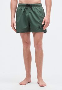 Zaļas peldbikses no viegla materiāla ar elastīgu jostu, kuru papildina aukliņa, un melnu drukātu logotipu.