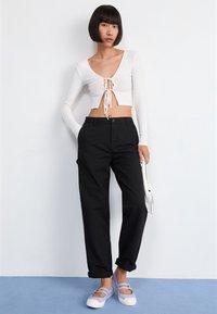 Biały prążkowany krótki top z wiązaniem z przodu, czarne luźne spodnie oraz lawendowo-białe sneakersy. Modelka stoi na jednolitym tle.