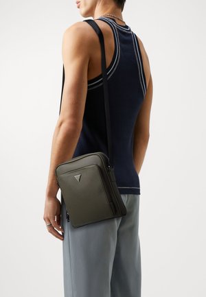 Personne portant un débardeur bleu marine et un pantalon gris, portant un petit sac bandoulière vert foncé avec un logo triangulaire sur le devant.