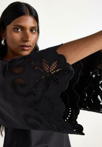 Svart plagg med intrikat spetsblommig broderi och scallopärmar. Mjuk textur med en flytande form och detaljerade utskärningar.