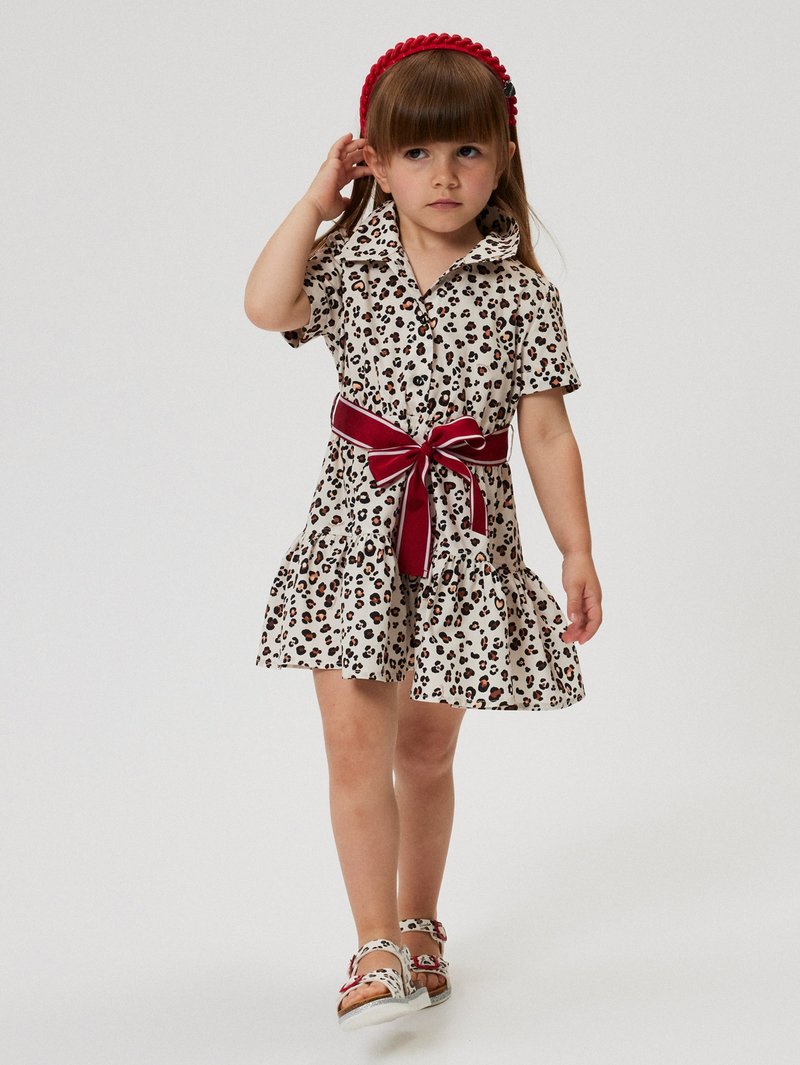 Leopard-print dress with short sleeves, a white base, ruffled hem, and a wide red belt. Paired with matching patterned sandals and a red headband.