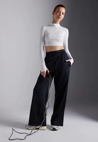 Weißes, kurzes Langarmoberteil mit halbem Reißverschluss, kombiniert mit lockeren schwarzen Hosen. Das Model hält eine Leine und trägt helle Sneakers.