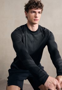 Črna dolgonoga športna majica z teksturiranim materialom in subtilnimi črtami na rokavih, v kombinaciji s črnimi šortsi, ki jih nosi sedeči model.