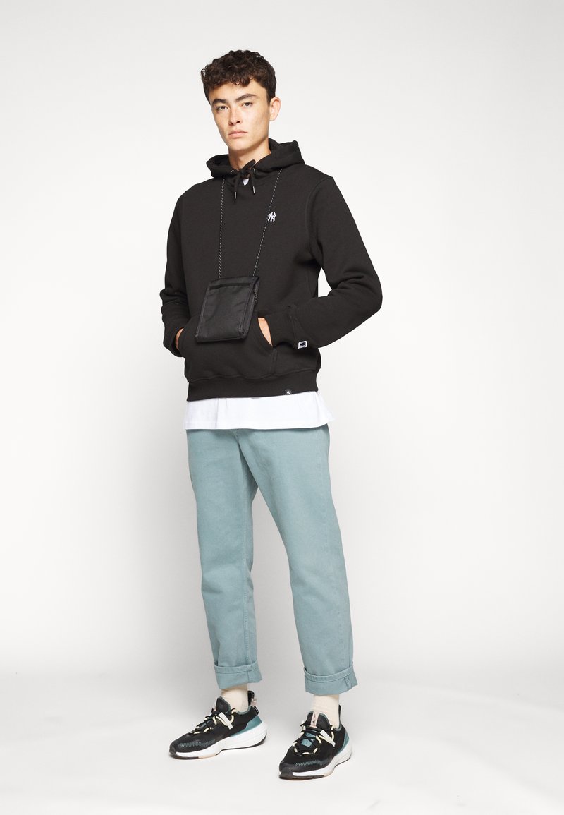 Zwarte hoodie met voorzak, turkooise taps toelopende broek en zwarte sneakers. Hoodie heeft een trekkoord en een logo. Casual outfit.