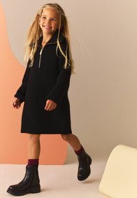Abito in maglia nera con colletto con zip, maniche lunghe e vestibilità rilassata. Abbinato a stivali neri sopra la caviglia e calze borgogna.