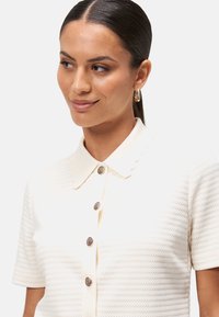 Kortærmet strikket polo-shirt i creme med et struktureret mønster, der har en spids krave og dekorative knapdetaljer i kontrasterende farver.