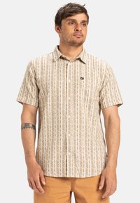 Camisa de botones de manga corta en color beige con patrones verticales marrones. Cuenta con un bolsillo y un pequeño logo en el pecho, fabricada con una tela ligera.