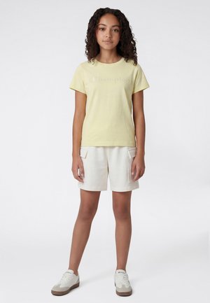 Joven de pie mirando hacia adelante, vestido con una camiseta Champion de color amarillo pálido, pantalones cortos cargo beige y zapatillas blancas sobre un fondo blanco.