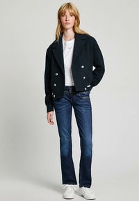 Casaco preto curto de abotoamento duplo com botões prateados, usado sobre uma camisa branca e combinado com jeans azuis de lavagem flare e sapatilhas brancas.