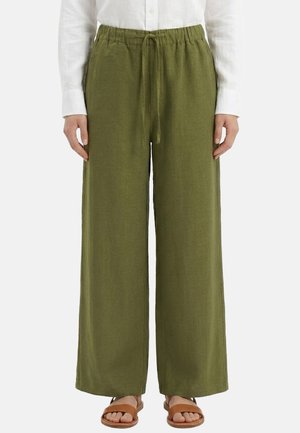 Donna che indossa pantaloni verde oliva a gamba larga con vita elasticizzata e coulisse, camicia bianca a maniche lunghe e sandali marroni aperti sulle dita.